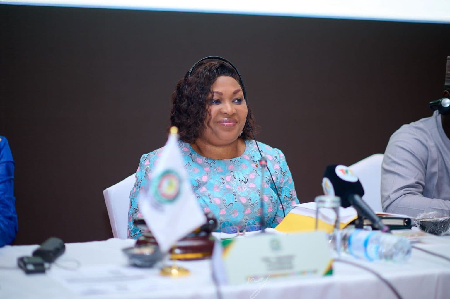 A woman seated at a conference table with an ECOWAS flag, wearing headphones during a parliamentary session