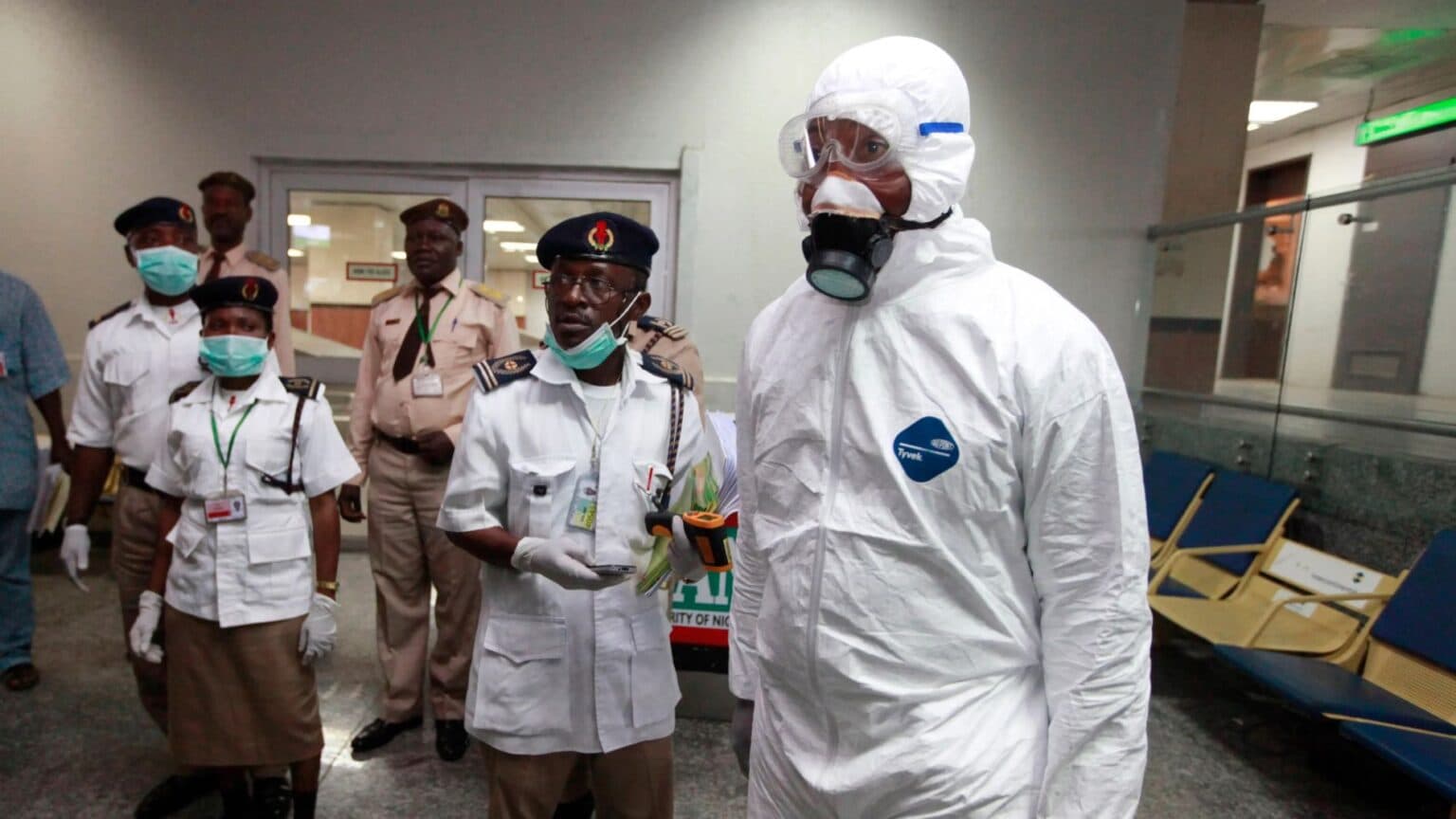 A health official in full protective suit and mask walking through an airport terminal alongside security personnel